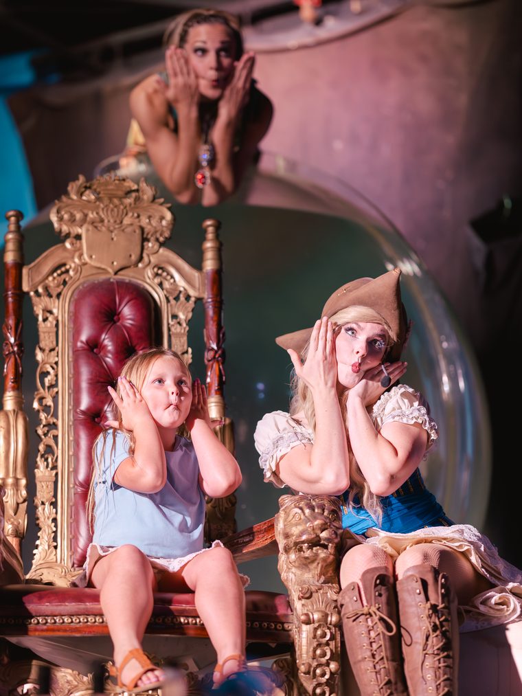 Two young guests blow kisses from a golden throne beside a costumed performer