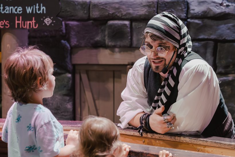 A bespectacled pirate leans down to talk with two small children at the rail