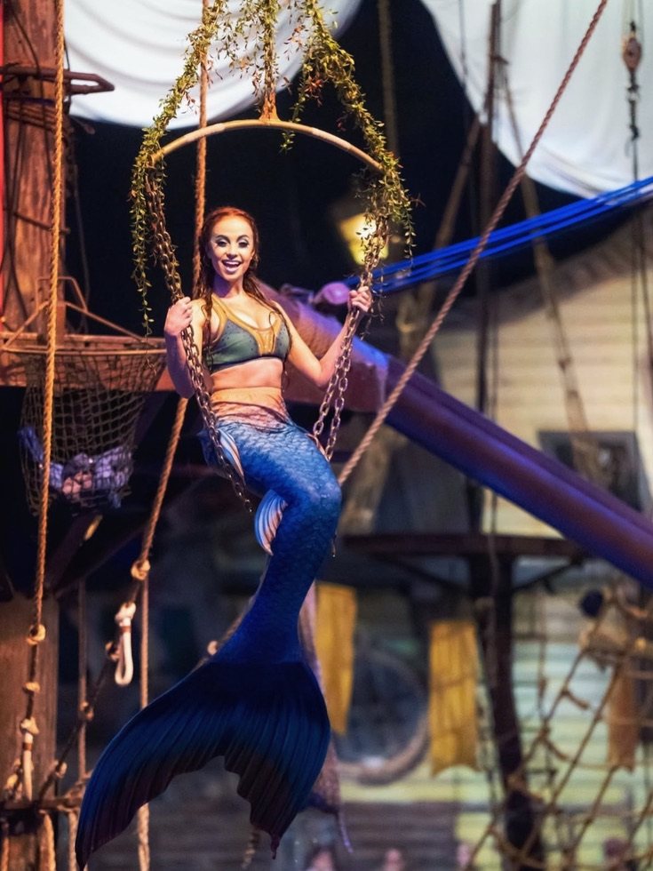 A mermaid performer poses in a suspended hoop above the deck